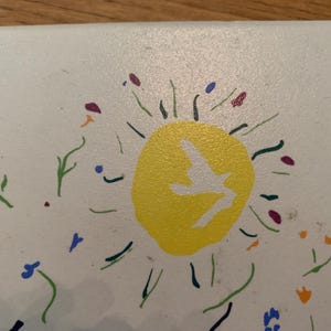 Pu&ograve; includere: Una silhouette di un uccello bianco che vola all'interno di un sole giallo con forme di fiori verdi, blu e viola intorno.