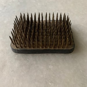 May include: A black and brown floral arranging tool with a square base and many metal pins.