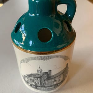 Könnte beinhalten: Kleine Keramikkrug mit tealgrünem Deckel und Henkel sowie weißem Sockel. Der Sockel zeigt eine Schwarz-Weiß-Illustration eines Gebäudes. Der Krug hat einen goldenen Rand und mehrere Löcher im Deckel.