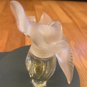 May include: A clear glass bottle of perfume with a frosted glass stopper shaped like a bird. The bottle is sitting on a dark green surface.