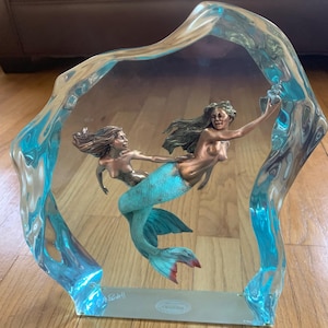 May include: A turquoise and bronze sculpture of two mermaids, encased in a clear blue resin block. The sculpture is signed "M. Findley" and "Edition".