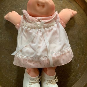 Könnte beinhalten: Eine Vintage-Cabbage-Patch-Kid-Puppe mit einem pfirsichfarbenen Gesicht und Gliedmaßen. Die Puppe trägt ein rosa Kleid mit weißem Overlay, Spitzensaum und weißer Schleife. Weiße Turnschuhe vervollständigen das Outfit.