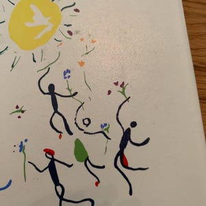 Pu&ograve; includere: Uno sfondo bianco con un sole giallo che contiene una colomba bianca. Ci sono quattro figure di bastoncino in nero, blu e rosso, che ballano intorno al sole. Le figure tengono fiori blu, rossi e verdi.