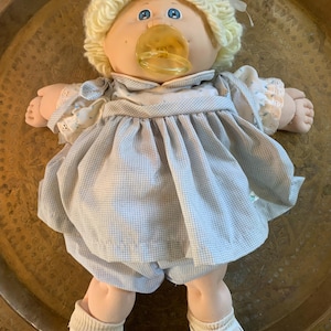 Pode incluir: Uma boneca Cabbage Patch Kid vintage com cabelo loiro de fio, olhos azuis e uma chupeta. A boneca usa um vestido xadrez azul e branco, calças combinando e sapatos brancos. A boneca está em uma superfície de cor de latão.