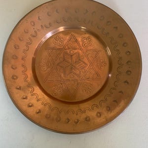 May include: A round copper plate featuring a central six-pointed star design composed of etched lines and small circular patterns. The outer rim is decorated with a repeating border of stamped semicircles and small square motifs in the metal.