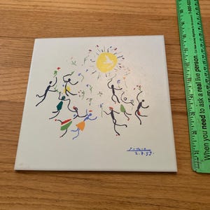 Pu&ograve; includere: Una piastrella di ceramica bianca con un'illustrazione colorata disegnata a mano di persone che ballano intorno a un sole giallo con una colomba bianca. La piastrella &egrave; firmata "2.7.59."