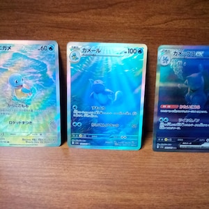 May include: Three Pokemon trading cards featuring Squirtle, Wartortle, and Blastoise. The cards have blue and turquoise backgrounds with Japanese text and character illustrations. The cards are displayed on a wooden surface.