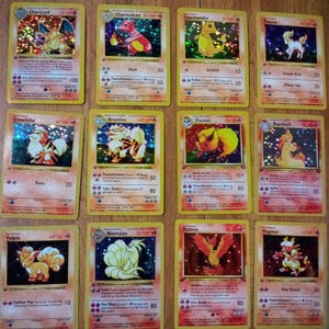 May include: A collection of twelve Pokémon trading cards, featuring various characters like Charizard, Charmeleon, and Charmander. The cards have a yellow border and include character names, HP, and attack details. The cards are displayed on a wooden surface.