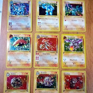 May include: A collection of nine holographic Pokémon trading cards arranged in a three-by-three grid. The cards feature various characters including Machamp, Machoke, Machop, Kabuto, Kabutops, Aerodactyl, Golem, Graveler, and Geodude with text.