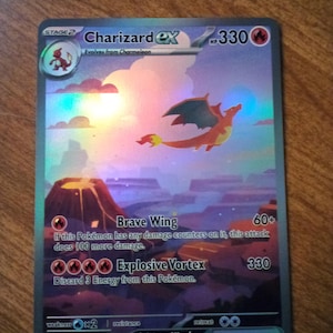 May include: A Charizard ex Pokémon trading card. The card features a holographic image of a flying orange dragon over a purple and brown landscape. The card has text that includes "Brave Wing" and "Explosive Vortex".