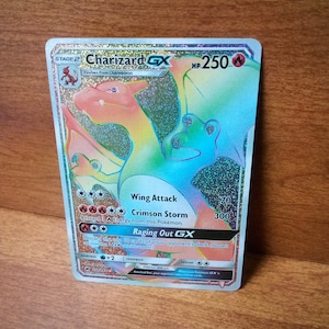 May include: A holographic Charizard GX trading card. The card features a colorful illustration of Charizard with the text "Charizard GX", "Wing Attack", "Crimson Storm", and "Raging Out GX". The card has a HP of 250.