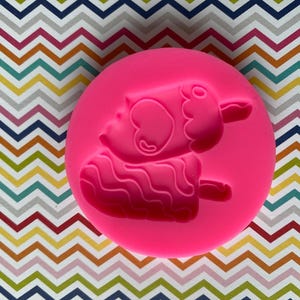 May include: A bright pink silicone mold featuring a cartoon sheep-shaped popsicle design. The mold is round and the sheep has a heart-shaped ear. The popsicle has a wavy texture and a stick. The background is a colorful chevron pattern.