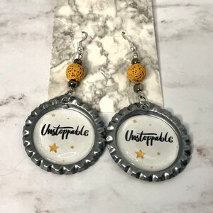May include: A pair of silver earrings with a yellow bead and a silver bottle cap charm that says "Unstoppable" with gold stars.
