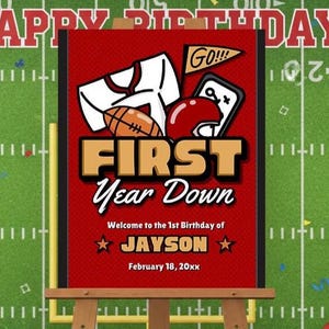 Peut inclure: Panneau d'anniversaire sur le thème du football, rouge et blanc, sur un chevalet. Le panneau indique "FIRST Year Down" avec des graphiques de football et le nom "JAYSON". Le fond est un terrain de football vert avec le texte "HAPPY BIRTHAY".
