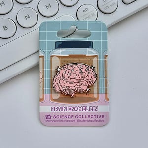 May include: A pink brain enamel pin on a backing card. The pin is shaped like a brain and is inside a jar illustration. The card has the text "BRAIN ENAMEL PIN" and "SCIENCE COLLECTIVE". A white keyboard is in the background.