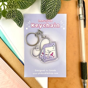 May include: A clear acrylic keychain featuring a tea bag design with the text "ImmuniTEA Keychain." The tea bag is light purple with pink and white details, attached to a silver keyring. The card reads "Designed in Canada" and "phdoodleshop.com."