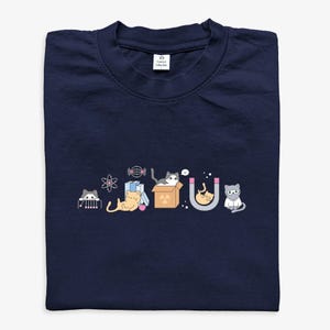 May include: Navy blue t-shirt with a science-themed illustration of cartoon cats interacting with scientific elements. The design includes an atom, books, a cat in a box, a magnet, and a cat wearing glasses. The tag reads "Science Collective".