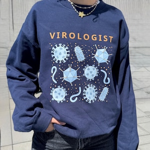 Op de afbeelding: Marineblauwe sweatshirt met het woord "VIROLOGIST" in oranje. Het ontwerp toont lichtblauwe illustraties van virussen en micro-organismen, waaronder geometrische vormen en stippenpatronen. Een casual kledingstuk.
