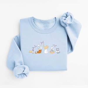 May include: A light blue sweatshirt featuring a whimsical illustration of cats in a laboratory setting. The design includes beakers, test tubes, and molecular structures, with various cats interacting with the scientific equipment. The sweatshirt has long sleeves and a crew neck.