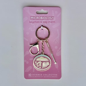 May include: A microbiology keychain and bag charm on a pink display card. The keychain features a rose gold ring, a lobster clasp, a small inoculating loop charm, and a round enamel charm depicting a petri dish with bacterial colonies.