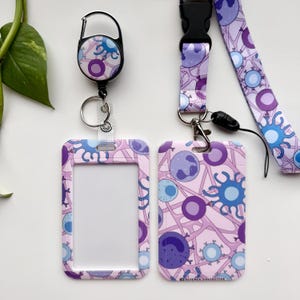 Könnte beinhalten: Ein hellrosa Schlüsselband-Set mit passendem Ausweishalter und einziehbarer Rolle. Das Set zeigt ein sich wiederholendes Muster aus blauen, violetten und weißen zellartigen Formen. Der Ausweishalter hat ein klares Fenster.