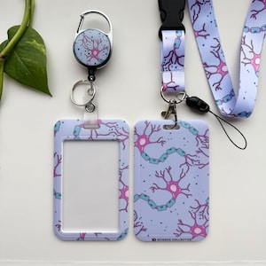 Può includere: Un set di cordino, porta badge e avvolgitore retrattile lilla chiaro. Il set presenta un motivo ripetuto di illustrazioni di neuroni rosa e blu. Il porta badge ha una finestra trasparente.