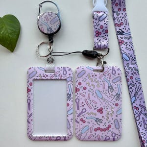 Könnte beinhalten: Ein hellviolettes Ausweishalter-Set mit einem sich wiederholenden Muster aus bunten Bakterien und Mikroorganismen. Das Set besteht aus einer vertikalen Ausweishülle aus Kunststoff, einem einziehbaren Ausweisjojo und einem passenden Schlüsselband.