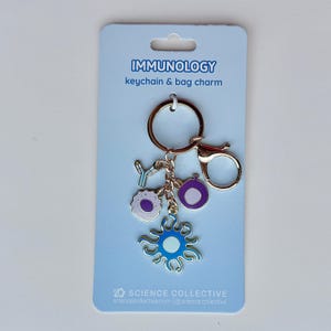 Peut inclure: Un porte-clés et breloque de sac immunologie sur une carte de présentation bleu clair. Le porte-clés comprend un anneau et un fermoir mousqueton dorés avec quatre breloques en émail : un anticorps en Y, une cellule violette texturée, une cellule violette et une cellule bleue en forme de soleil.