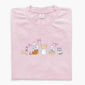 May include: A pale pink t-shirt with a science-themed illustration. The design shows cartoon cats with laboratory equipment like beakers and test tubes, alongside molecular structures. The cats are in different poses, giving a playful feel to the design.