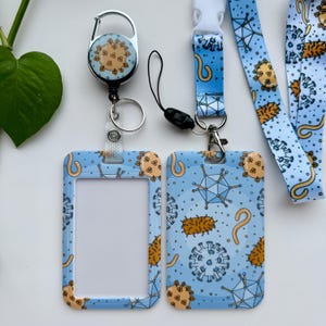 Peut inclure: Ensemble de lanière et porte-badge bleu clair avec un motif répété d'illustrations de virus orange et bleu. Le porte-badge a une fenêtre transparente pour une carte d'identité. Un enrouleur rétractable avec un design similaire est également inclus.