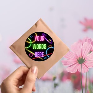 Puede incluir: Un sobre de papel marr&oacute;n con una pegatina redonda con luces de ne&oacute;n rosa, azul, verde y amarillo. La pegatina dice "YOUR WORDS HERE" en texto negro.