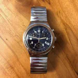 May include: A silver-tone Swatch chronograph watch with a dark blue dial, white Arabic numerals, three sub-dials, and a date window at the four o'clock position. The watch features a metallic link bracelet and a tachymeter scale on the bezel.