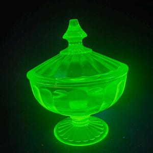 May include: A glowing neon green glass candy dish with a matching lid. The bowl features a ribbed pattern along the sides and sits on a circular pedestal base. The lid has a pointed finial top and matching vertical ribbing, emitting a bright light.