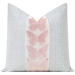 May include: A decorative square pillow with a light gray textured fabric. A vertical strip of white fabric runs down the center, adorned with four pink butterfly appliqués. The pillow is designed for home decor.