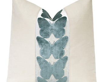 May include: A square throw pillow with a cream-colored fabric base. A vertical central panel features a series of four stacked, teal-colored velvet butterfly appliques. The pillow has a subtle, fine-textured weave throughout the cream sections.