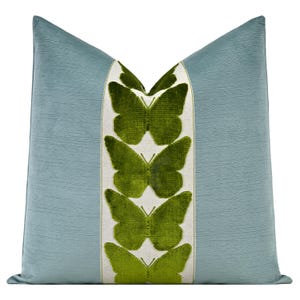 May include: A light blue cushion with a textured surface. A central panel features green butterfly motifs on a white background. The cushion has a soft, plush appearance and a classic shape, suitable for a sofa or bed.