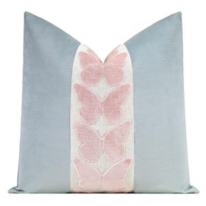 May include: A decorative throw pillow with a light blue background and a central panel featuring four pink butterfly appliqués. The butterflies are embroidered with a textured design. The pillow has a soft, velvety appearance.