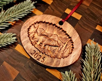 May include: Oval wooden ornament featuring a carved bear and mountain landscape, with the year 2025 etched below. A red ribbon is attached for hanging. The ornament is surrounded by evergreen branches.