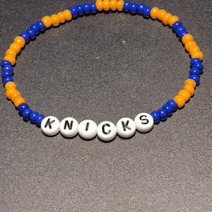 May include: A beaded bracelet featuring the word "KNICKS" spelled out in white letter beads. The bracelet alternates between blue and orange beads, the colors of the New York Knicks basketball team. The bracelet is on a dark gray surface.