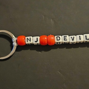 May include: A red and white beaded keyring with the letters "NJ DEVILS" spelled out. The keyring has a silver metal ring and a silver metal clasp.