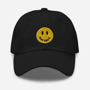 Può includere: Cappellino da baseball nero con una faccia sorridente gialla ricamata sul davanti. La faccia sorridente ha il testo "Don't fake it" scritto all'interno.