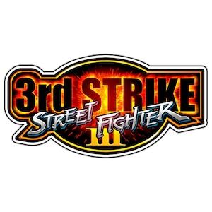 May include: A sticker featuring the "3rd STRIKE Street Fighter III" logo. The logo has a black outline with a yellow and orange gradient background. The text is in white and silver.