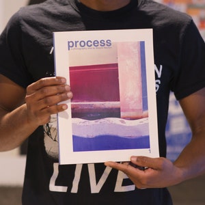 May include: A person holding a magazine titled "process" with a photography by Daphne Brickson. The cover features a photograph of a pink and blue wall with a blue and white wave pattern at the bottom.