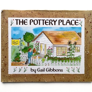 May include: A rectangular book titled The Pottery Place by Gail Gibbons. The cover features a watercolor illustration of a white house with a brown roof, a person holding a pot, a cat, and geese behind a white picket fence, all on a speckled tan border.