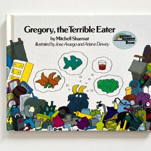 May include: A children's book cover titled "Gregory, the Terrible Eater" by Mitchell Sharmat, illustrated by Jose Aruego and Ariane Dewey. The cover features a cartoon goat surrounded by various objects, including a fish, a carrot, a glass of juice, and a leafy green vegetable.