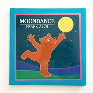 May include: A blue book cover with a pink border. The title "MOONDANCE" is in white text. Below the title is "FRANK ASCH" in white text. The cover features a brown bear dancing with a yellow moon in the background.
