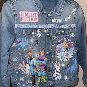 Puede incluir: Chaqueta vaquera azul claro con parche de "Edición Limitada", ribete de lentejuelas y gráficos de personajes de Frozen. Adornada con pedrería y detalles de copos de nieve. Incluye la frase "Let it go".