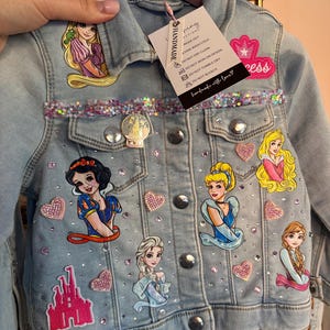 Puede incluir: Chaqueta vaquera azul claro decorada con parches de princesas, pedrería y adornos en forma de corazón. Presenta imágenes de princesas de Disney, un castillo y la palabra "Princess". Incluye una etiqueta que dice "HANDMADE".