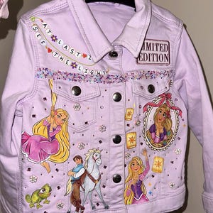 Może przedstawiać: Jasnofioletowa kurtka dżinsowa z ilustracjami księżniczki, konia i lampionów. Kurtka ma napis "AT LAST I SEE THE LIGHT" i "LIMITED EDITION". Ozdobiona cekinami i dżetami.