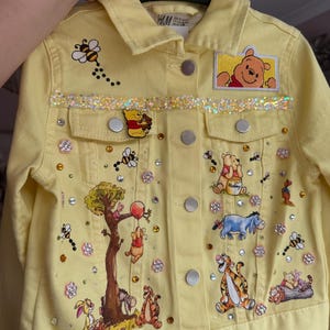 May include: Pale yellow denim jacket decorated with Winnie the Pooh characters, rhinestones, and bee motifs. Features a glitter trim and character patches. Includes a button-up front and two chest pockets.